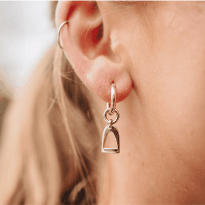 Equestrian Earrings in polished silver-tone metal, featuring a small stirrup charm suspended from a hinged huggie hoop.