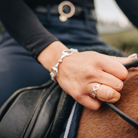 Photo of a girl riding a horse holding the reins in her hand. On her finger is a silver horse bit snaffle ring and in the background on her wrist she is wearing a horse bit snaffle curb chain bracelet. Both made by Crawford Hill Equestrian Jewellery.